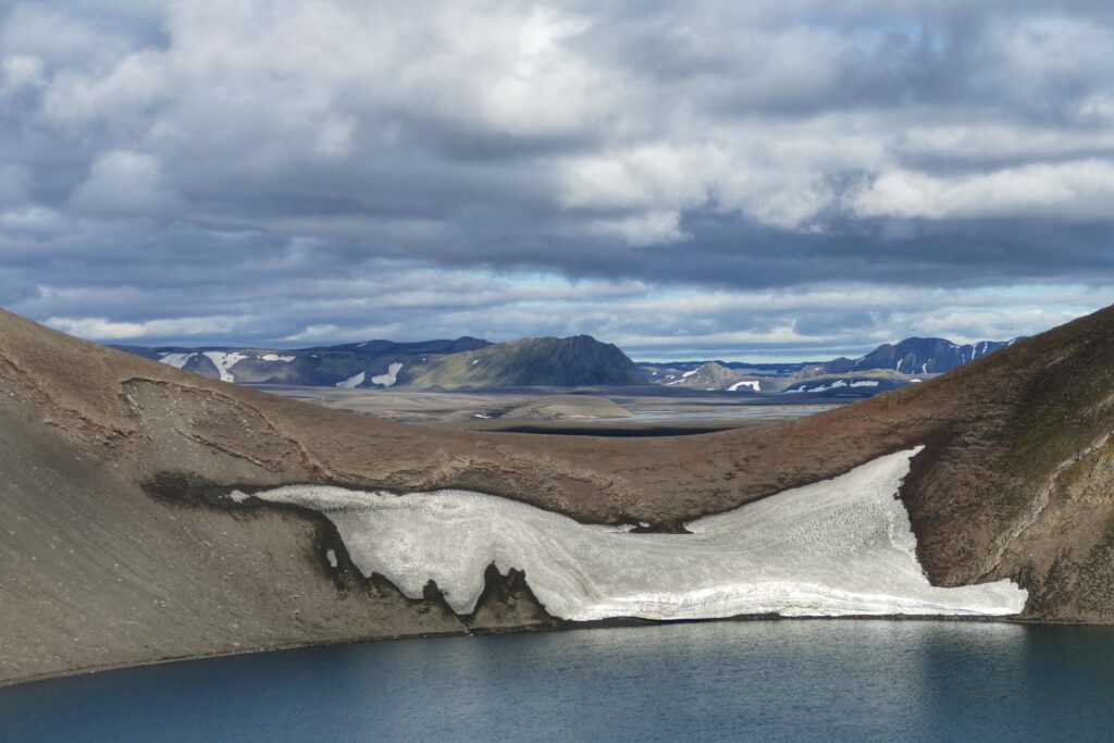 sopečné jezero s trochou sněhu a v dálce hory a zatažená obloha