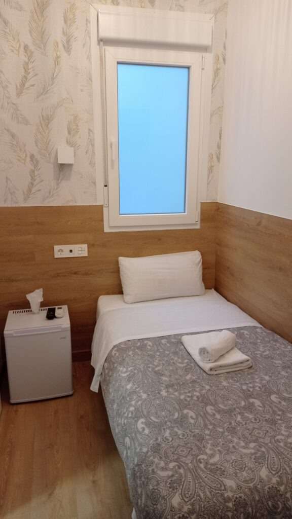Hotelový pokoj s jednou postelí, malou ledničkou a oknem
